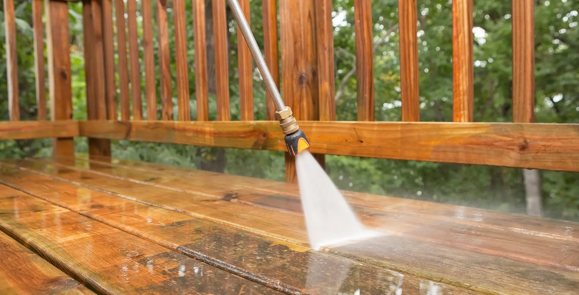 A person using a power washer on the side of a wooden deck.